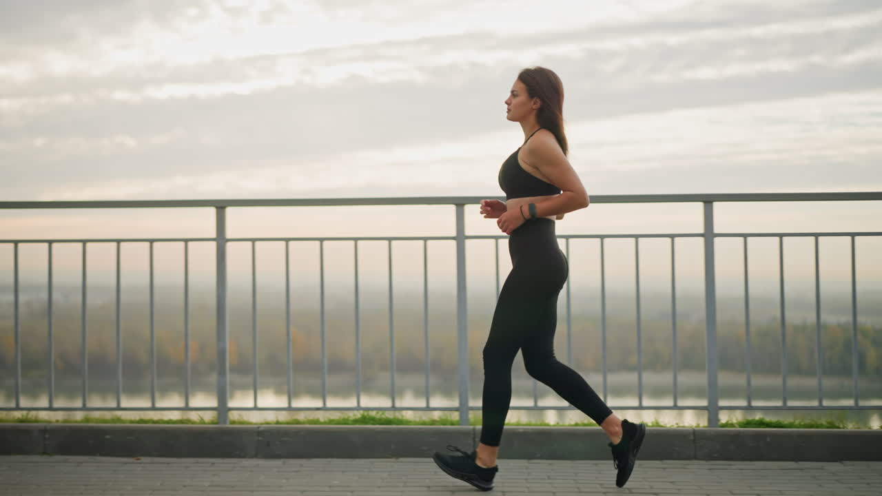 Woman in black top and leggings with sneakers running along road path beside iron fence, enjoying her fitness routine outdoors, with a scenic view of nature and distant landscape
