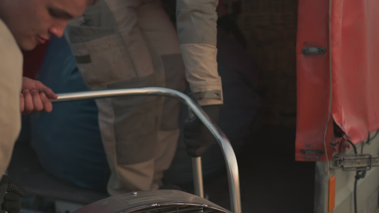 close up of adults stowing engine fan assembly into vehicle trunk at dusk, hands guiding motor grill into cargo space against blurred farm field and soft sky background