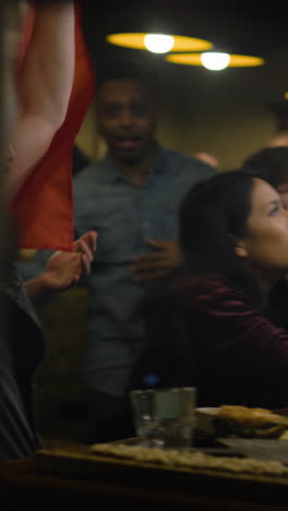 Vertical-shot-of-friends-cheering-for-soccer-team-playing-in-world-league-tournament.-Cheerful-sports-fans-applaud-and-shout-watching-football-match-live-broadcast-in-bar-or-sports-pub-at-night.