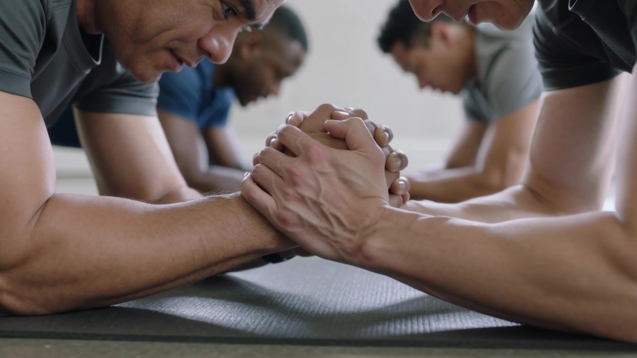 Coach cueing two men in short sleeves holding partner forearm plank on dark mats building strength