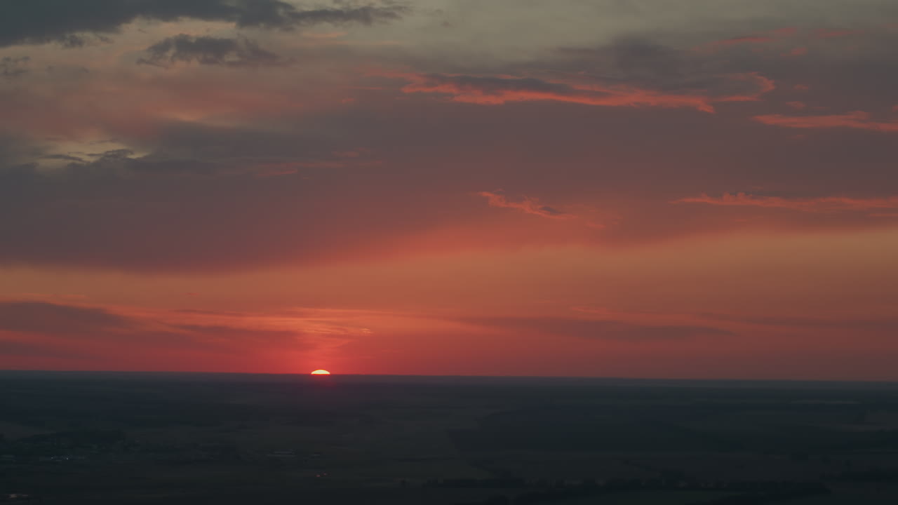 Rich red and orange evening sunset sky with dramatic dark clouds hovering above flat fields horizon, sun half dipping behind distant fields with subtle lens flare and pastel glow