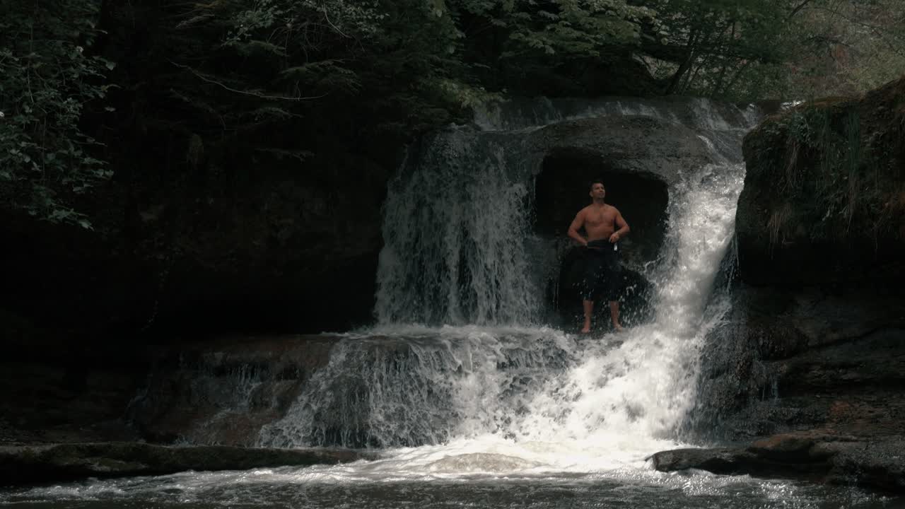 A man stands by a waterfall in an epic landscape. He is shirtless and enjoying nature. It's raining. Shot in an epic forest in slow motion.