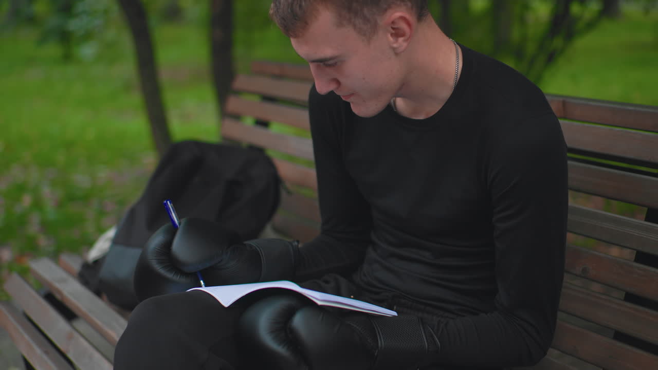 Jovem boxeador lendo um caderno em um banco de parque, com luvas descansando no colo, estudando táticas e anotações, mochila ao lado do banco, olhar concentrado, concentração de treinador, folhas de outono, calmaria urbana, sessão de planejamento