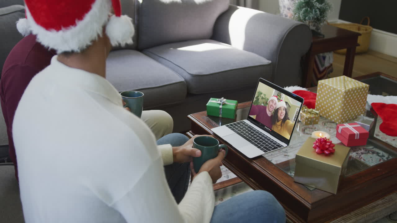 padre y hijo biraciales usando una computadora portátil para una videollamada de navidad con una pareja sonriente en la pantalla