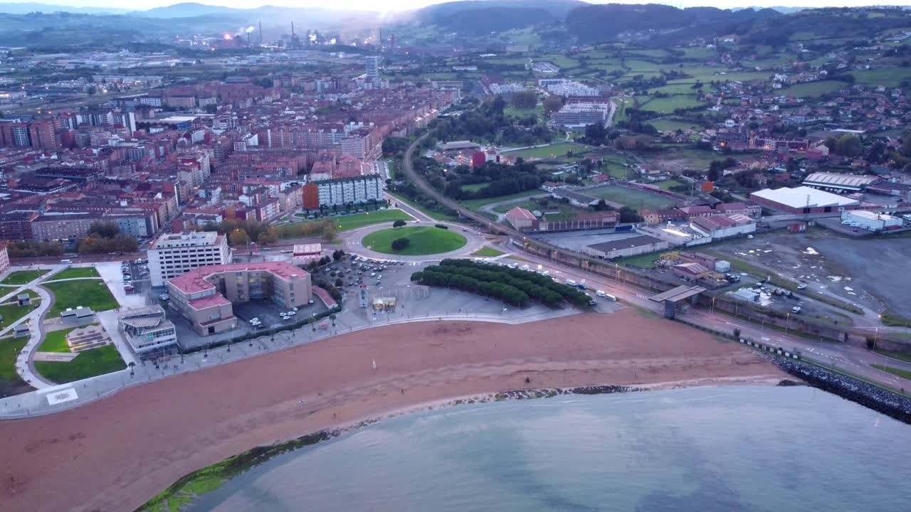 Vista aerea della città di Gijon nel nord della Spagna, con centro città commerciale e porto, spiaggia sabbiosa e area residenziale; fabbrica fumosa sullo sfondo con emissioni di gas inquinanti.