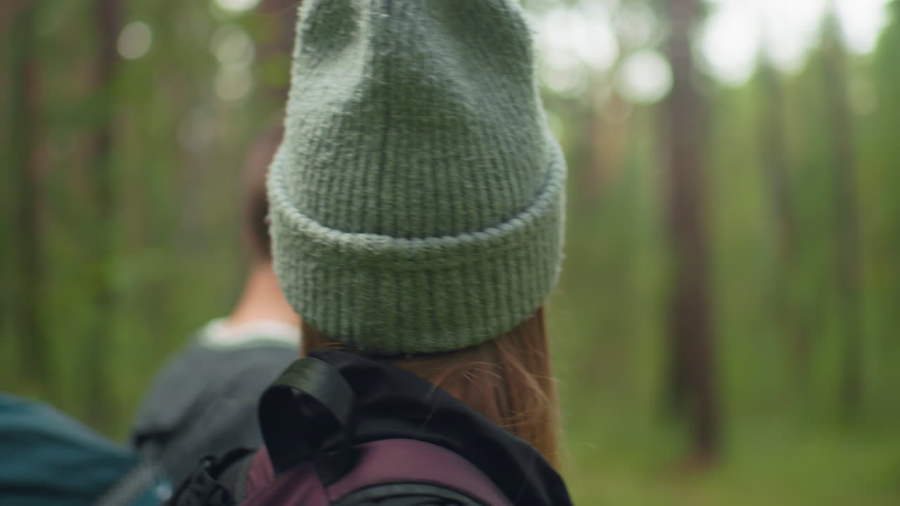 Close up of young woman in beanie and jacket standing in forest, appearing deep in thought as she looks around, with male companion slightly blurred in background