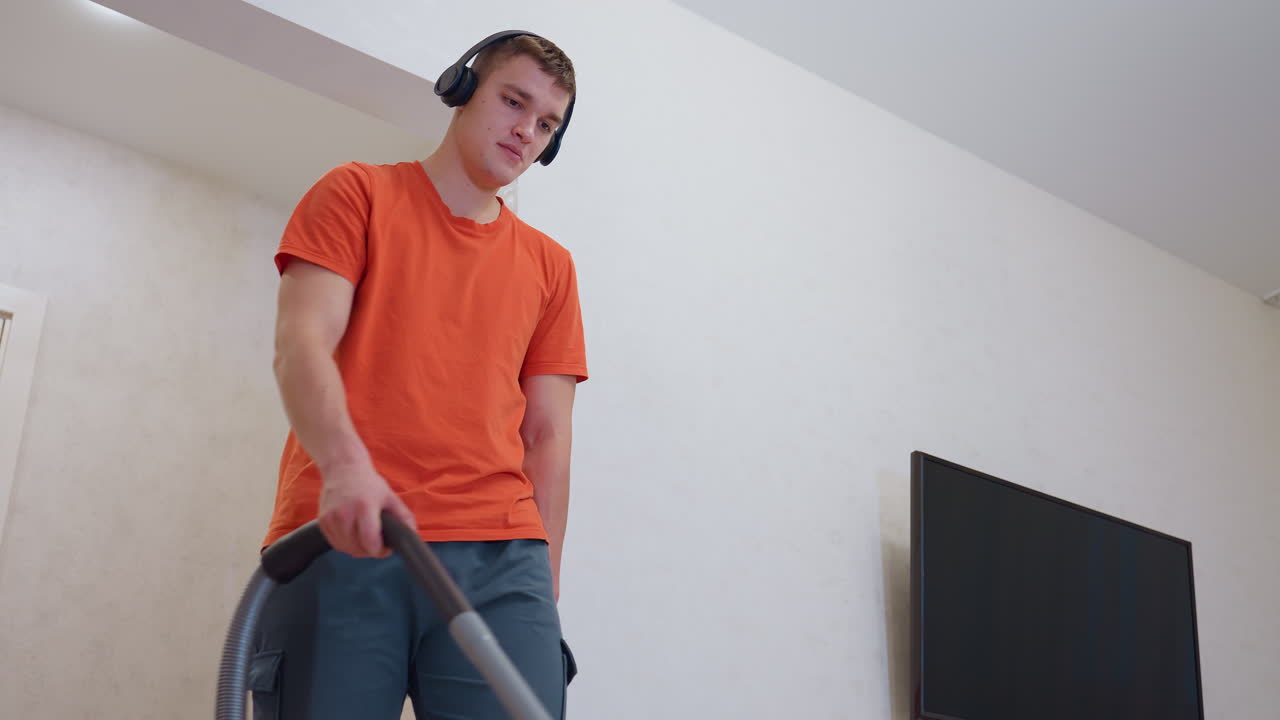 Young man in orange shirt wearing black headphones vacuuming room with focused expression while standing near wall and television, engaging in cleaning routine while listening to music indoors
