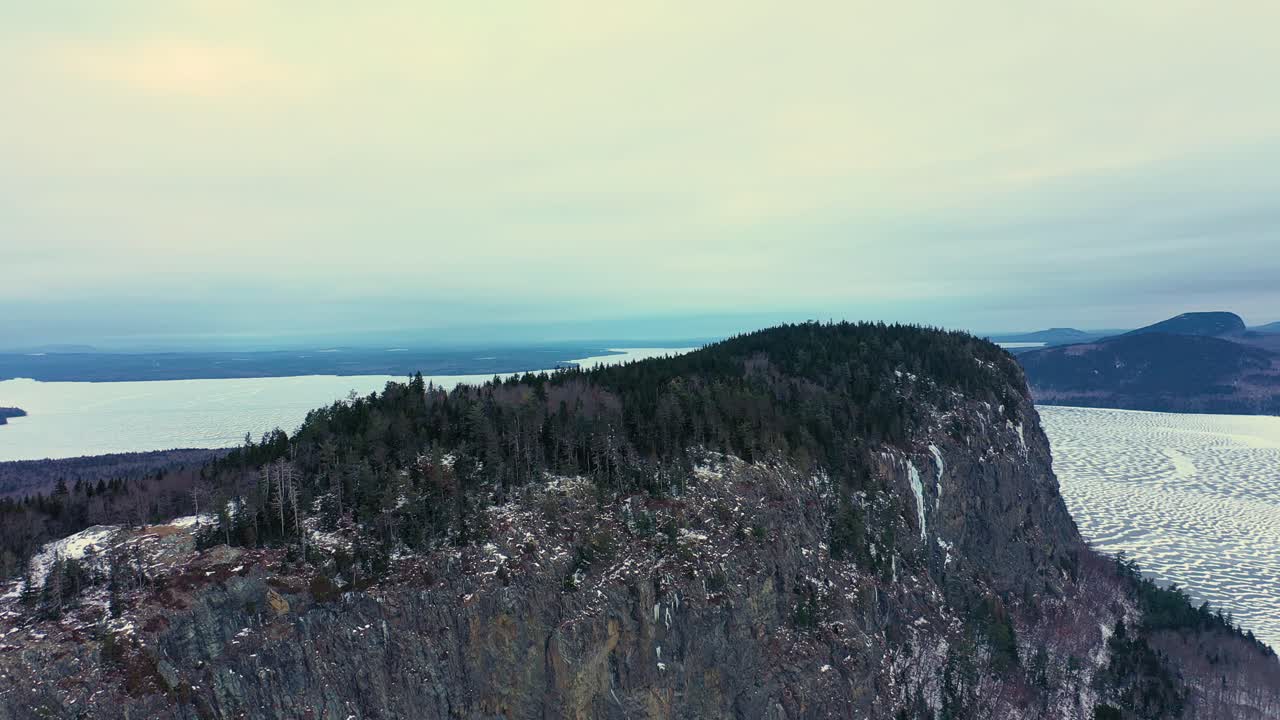 volando hacia atrás y hacia abajo para revelar la totalidad de una montaña aislada que se eleva desde un lago congelado con un acantilado empinado