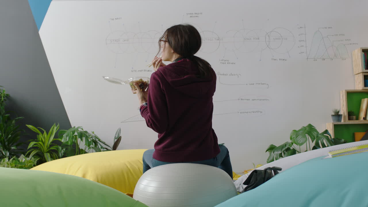 feliz mujer caucásica estudiante lluvia de ideas disfrutando de la pausa del almuerzo comiendo sentada en la pelota de ejercicio revisando mensajes de redes sociales en el teléfono inteligente en el lugar de trabajo moderno de la oficina