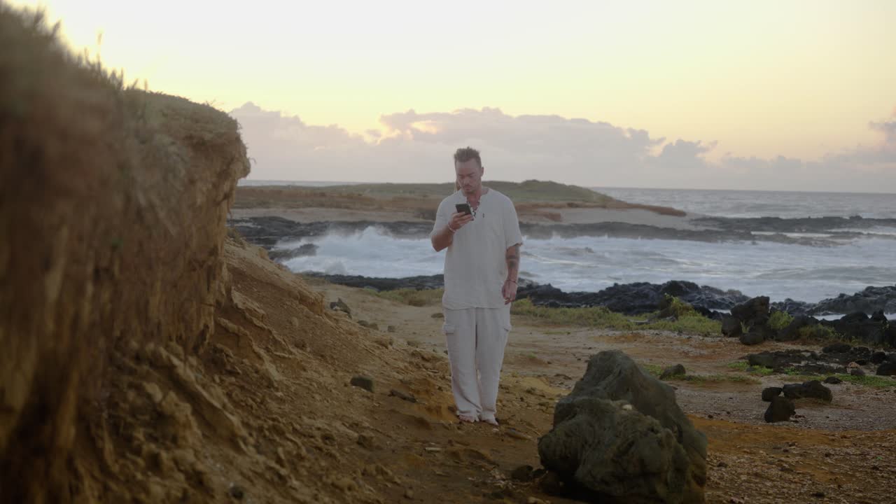 A man in white clothing walks toward the camera on a rugged coastal path, checking his smartphone as the ocean waves roll in under soft morning light