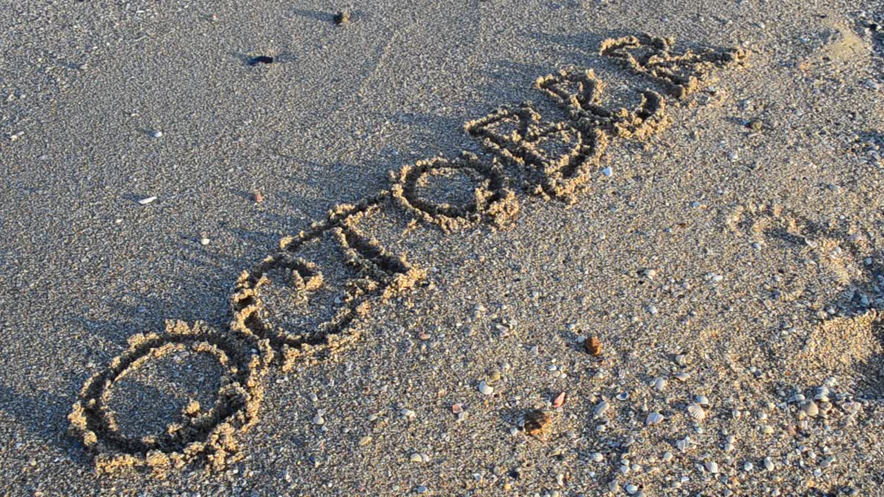 Inschrift Oktober im Sand. Die Inschrift wird von einer Meereswelle weggespült. Aufnahmen am Strand.