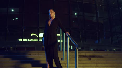 young woman stands on the stairway of a glass building in the city at night in a black dress