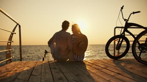 a happy couple, a guy and a girl, are sitting on the shore of a beach that is covered with boards and looking at the sunrise on the sea, their bike is located near them. happy date on the beach near the sea at sunrise