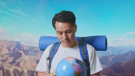 close up of asian male hiker with mountaineering backpack holding world globe in his hands and smiling to camera while traveling at the top of mountain