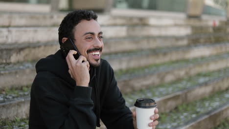 young arabic man with dark curly hair and beard in black hoodie sitting on stairs outside