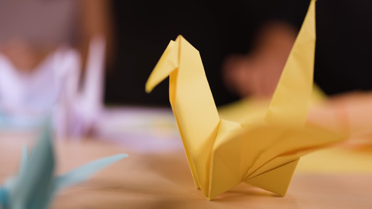 Una gru di carta gialla è appoggiata su un tavolo mentre delle mani piegano origami sullo sfondo