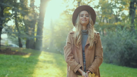 cheerful stylish caucasian young woman wearing a hat and coat having fun in the park, throwing autumn yellow leaves and looking at camera