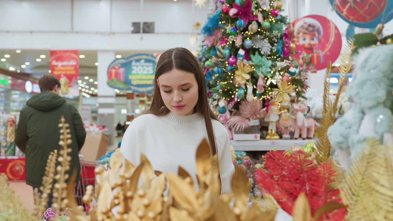 Woman in decor store holding and admiring a brown decorative ornament with a smile, surrounded by vibrant holiday decorations and other shoppers in the background
