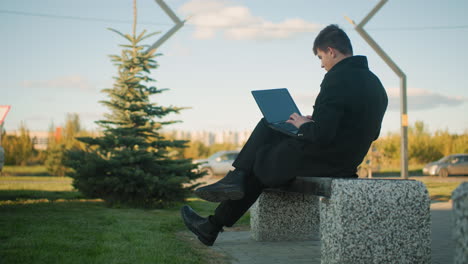 side view of man seated outdoors operating laptop with leg crossed, surrounded by greenery and urban structures, background includes parked cars and another car parking