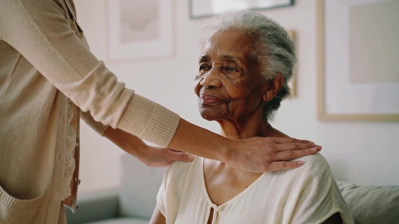 Reaching caregiver placing hands on senior in light blouse at home smoothing shoulders and calming