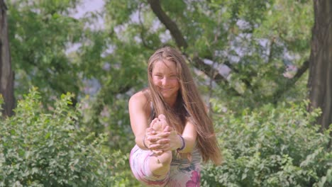 Front-view-of-young-girl-smiles-at-camera-while-stretching-leg-in-nature,-slomo