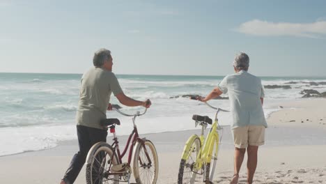 back view of hispanic senior couple walking with bikes on beach at sunset