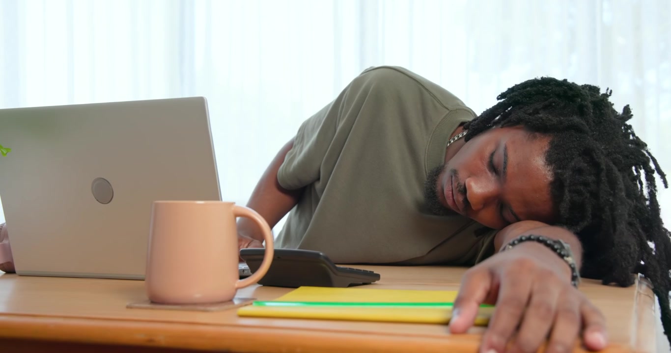 Dreadlocked man yawning rubbing eyes reaching notebooks at laptop desk dozing off