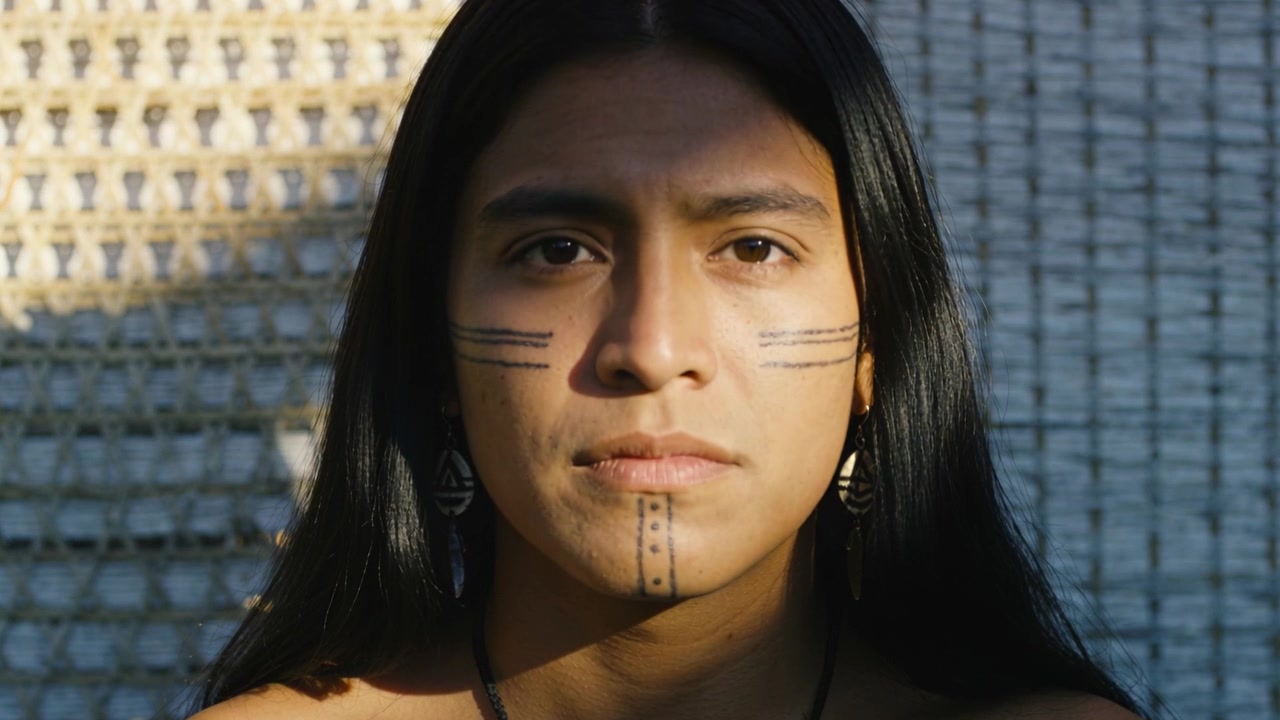 Uomo indigeno seduto che sbatte le palpebre, con la luce del sole che si sposta sulla fronte, vicino a un tappeto intrecciato con pittura e collana.