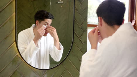 focused biracial man applying under eye masks looking in bathroom mirror, slow motion