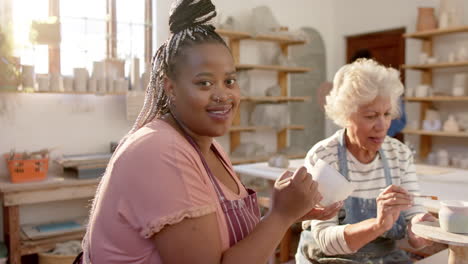 two diverse female potters glazing clay jug and smiling in pottery studio, slow motion