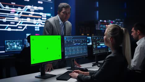 female specialist works on green screen computer talks with project manager in big infrastructure control room. monitoring room team of professionals use computers showing charts, information