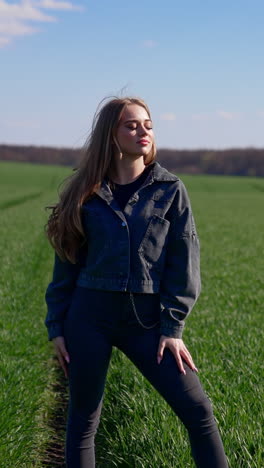 attractive girl posing on camera outdoors. beautiful young model in tight pants and cotton jacket with fluttering hair looking on camera on green field background. vertical video