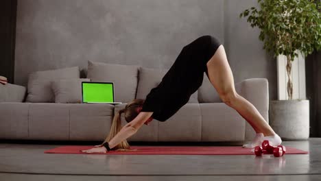 Mujer-Caucásica-Y-Rubia-Con-Ropa-Deportiva-Negra-Entrenando-En-Casa-Sobre-Una-Alfombra-Frente-Al-Sofá-Gris-Y-Estirando-Los-Músculos-De-Las-Piernas,-Sentada-En-El-Suelo,-Inclinando-El-Torso-Hacia-Adelante.-Habitación-Interior-Tipo-Loft.-Mancuernas-En-El-Suelo-Cerca-De-La-Colchoneta