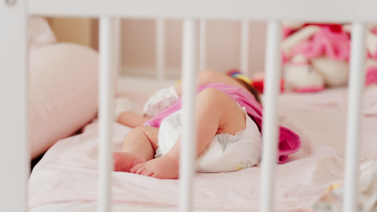 Close up of a baby's legs and diaper as the infant rests in a crib, seen through the crib bars