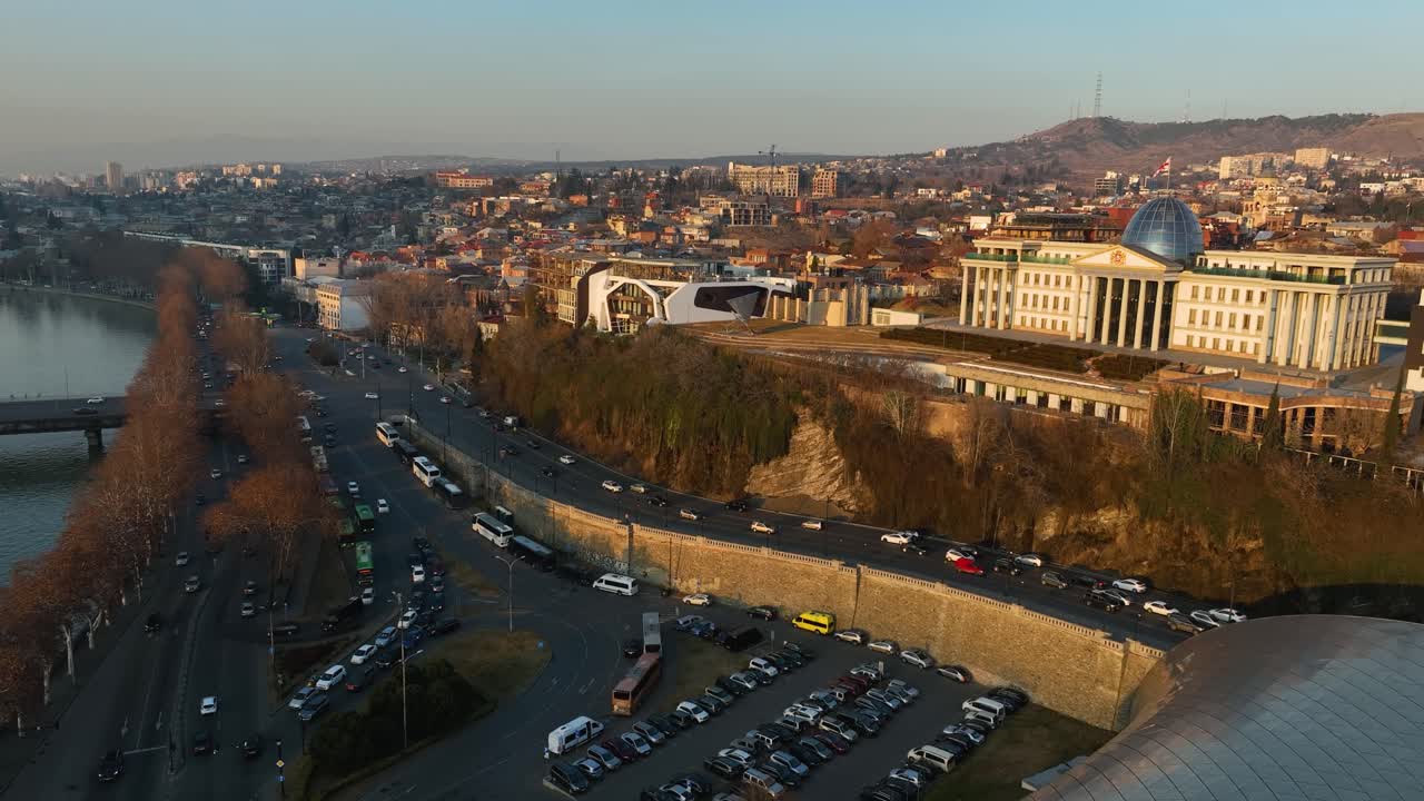 A stunning aerial view of Tbilisi’s modern riverfront featuring the tubular Rike Park Concert Hall surrounded by historic city architecture and autumn colors