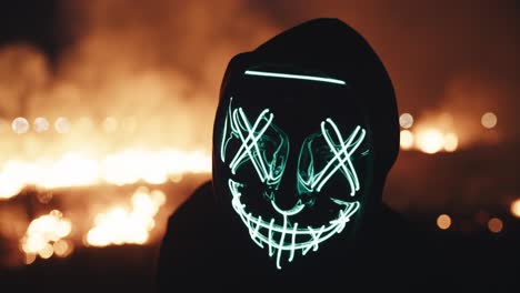 person in a glowing light-up mask at night near a fire