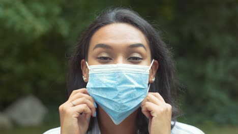 Retrato-De-Una-Linda-Mujer-Latina-Poniéndose-Una-Mascarilla-Médica-Con-Ambas-Manos-Mientras-Se-Pone-El-Pelo-Detrás-De-Las-Orejas,-Afuera-En-El-Parque