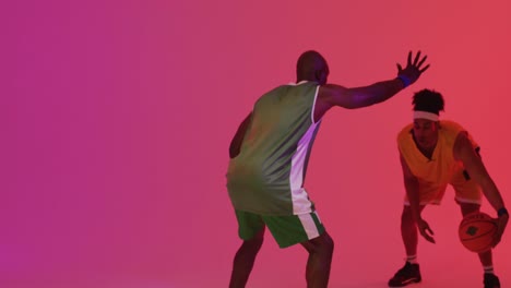 Video-of-two-diverse-male-basketball-players-playing-with-ball-on-pink-background