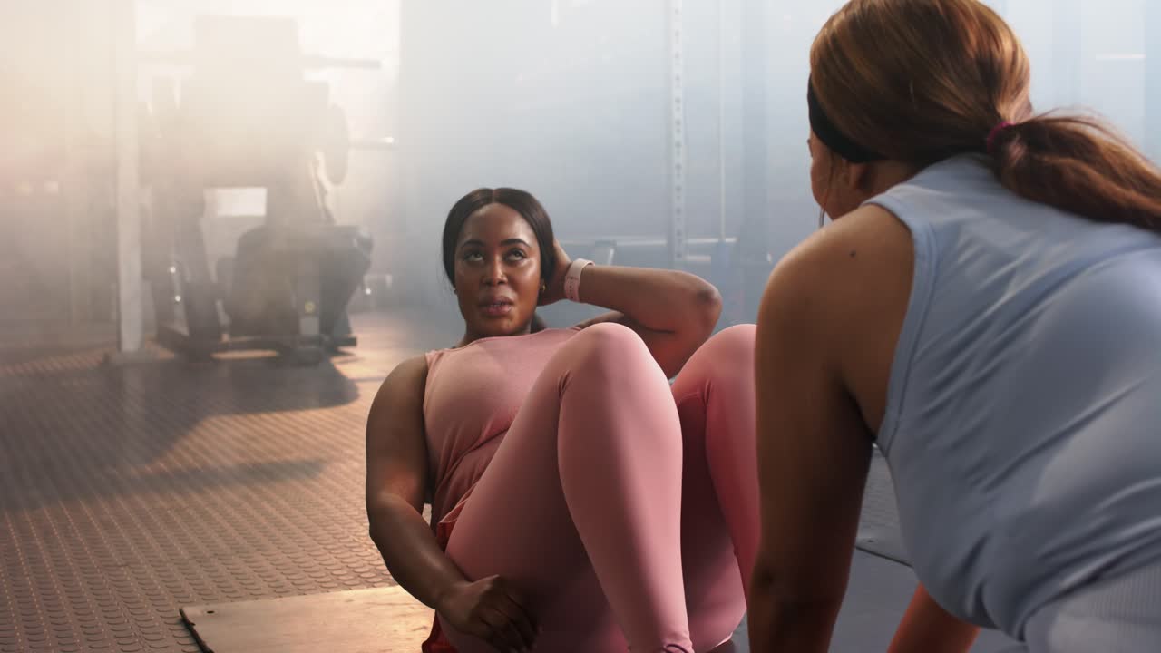 Diverse female workout partners doing sit-ups on mat in gym, partner bracing ankles starting reps