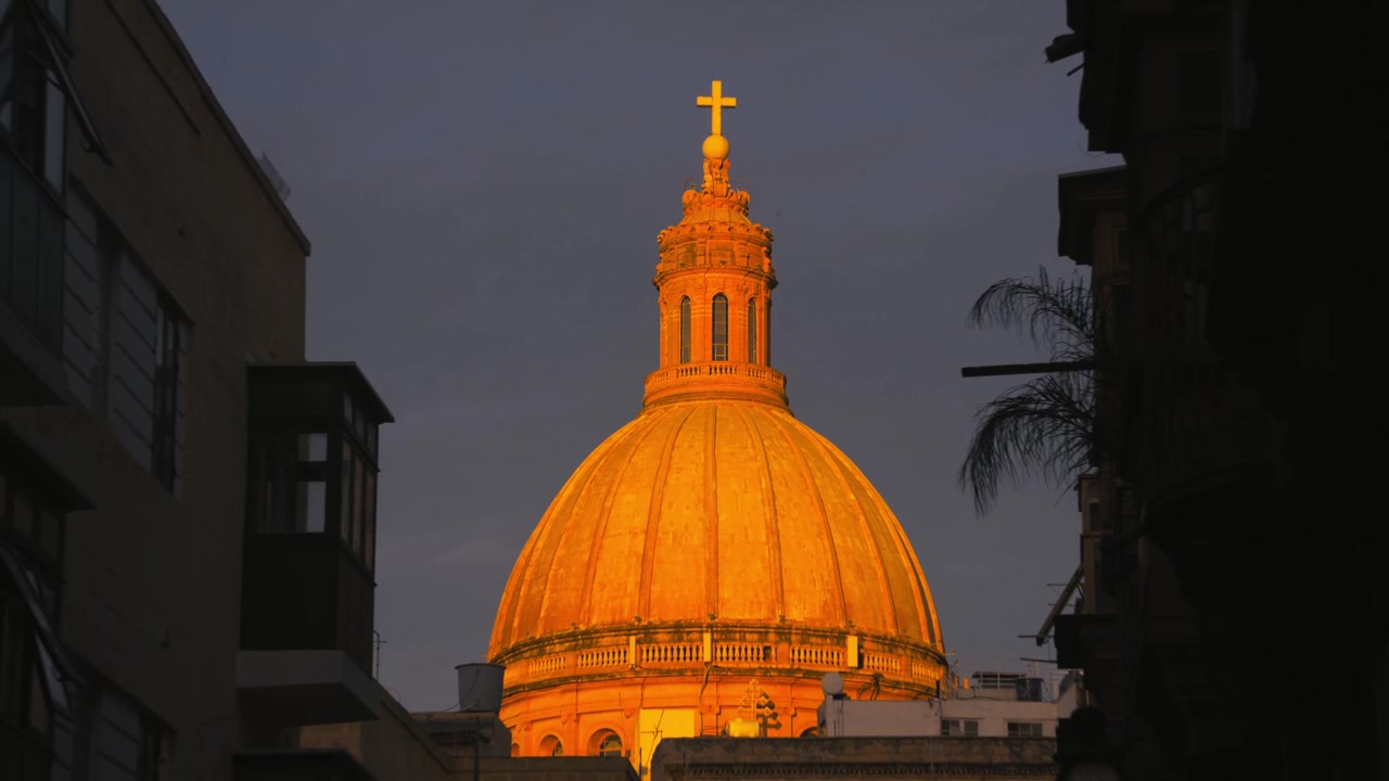 L'iconica cupola della Cattedrale di Nostra Signora del Monte Carmelo a La Valletta si illumina di una calda luce arancione, incorniciata dagli edifici della città al tramonto a Malta, Madonna tal-Karmnu.