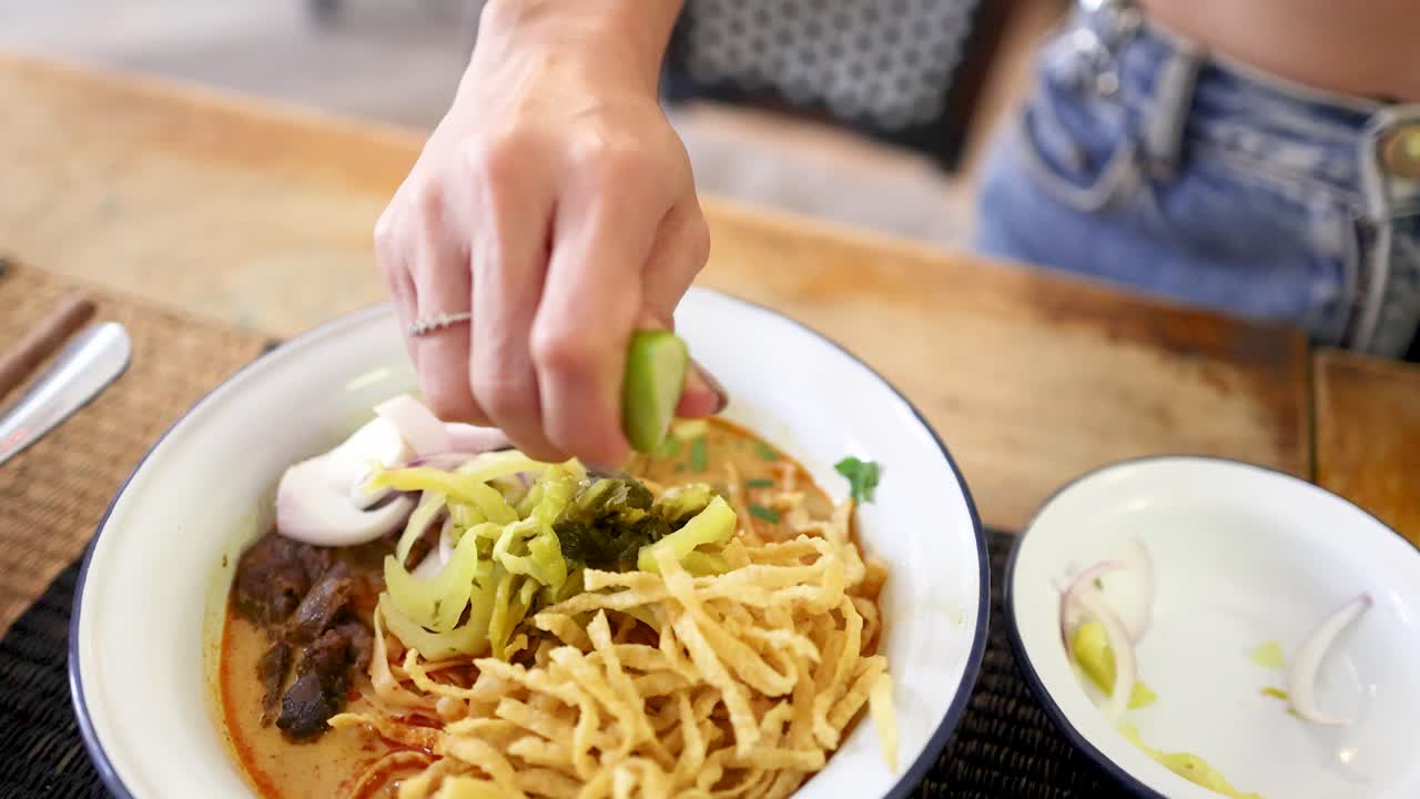 A hand squeezes fresh lime over a bowl of Northern Thai beef curry noodles at a restaurant table in Bangkok, under natural daylight with a close-up perspective