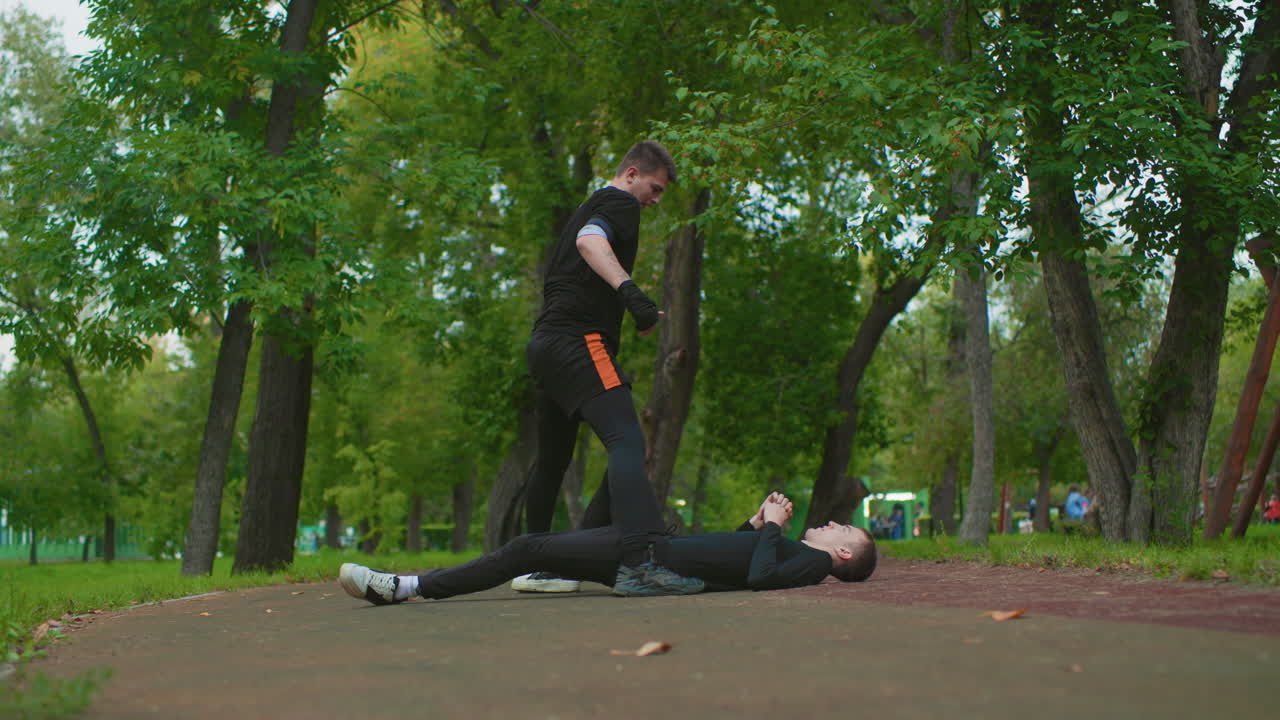 white male fighters reconciling in leafy park after intense training, handshake and calm discussion between partners, coach nodding as duo walk off pathway, camaraderie and sportsmanship replacing