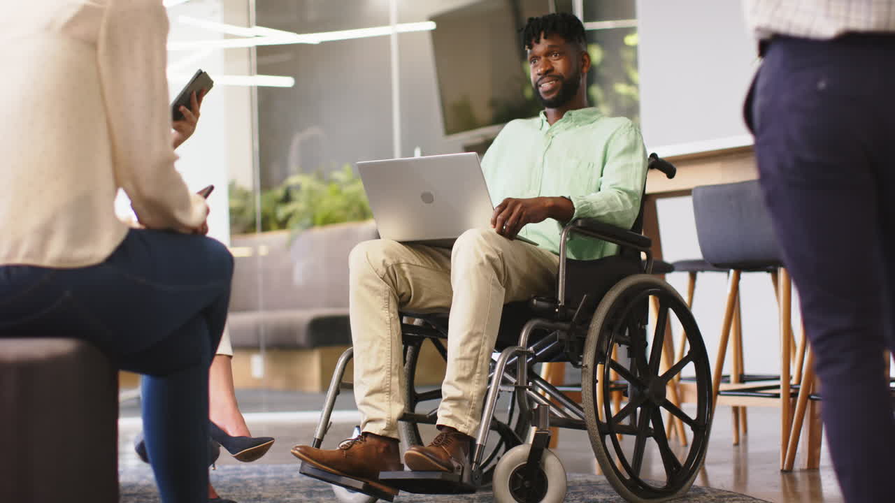 In office, man in wheelchair using laptop, engaging in group discussion