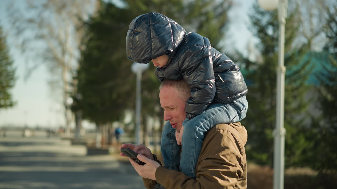 una vista lateral un hombre llevando a su hijo en el hombro centrado en su teléfono mientras lo opera, con una vista borrosa de árboles y personas en el fondo