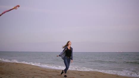 Happy-young-woman-with-long-hair-releasing-colorful-kite-on-the-beach-in-the-evening-during-sunset-and-running-with-it