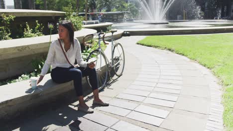 mujer afroamericana usando un teléfono inteligente bebiendo café en un parque de la ciudad