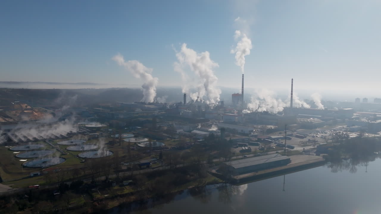 Veduta aerea dall'alto del sito industriale di Štětí che mostra un'intensa attività di vapore da più ciminiere, complessi edifici produttivi estesi e bacini circolari sotto un cielo sereno.