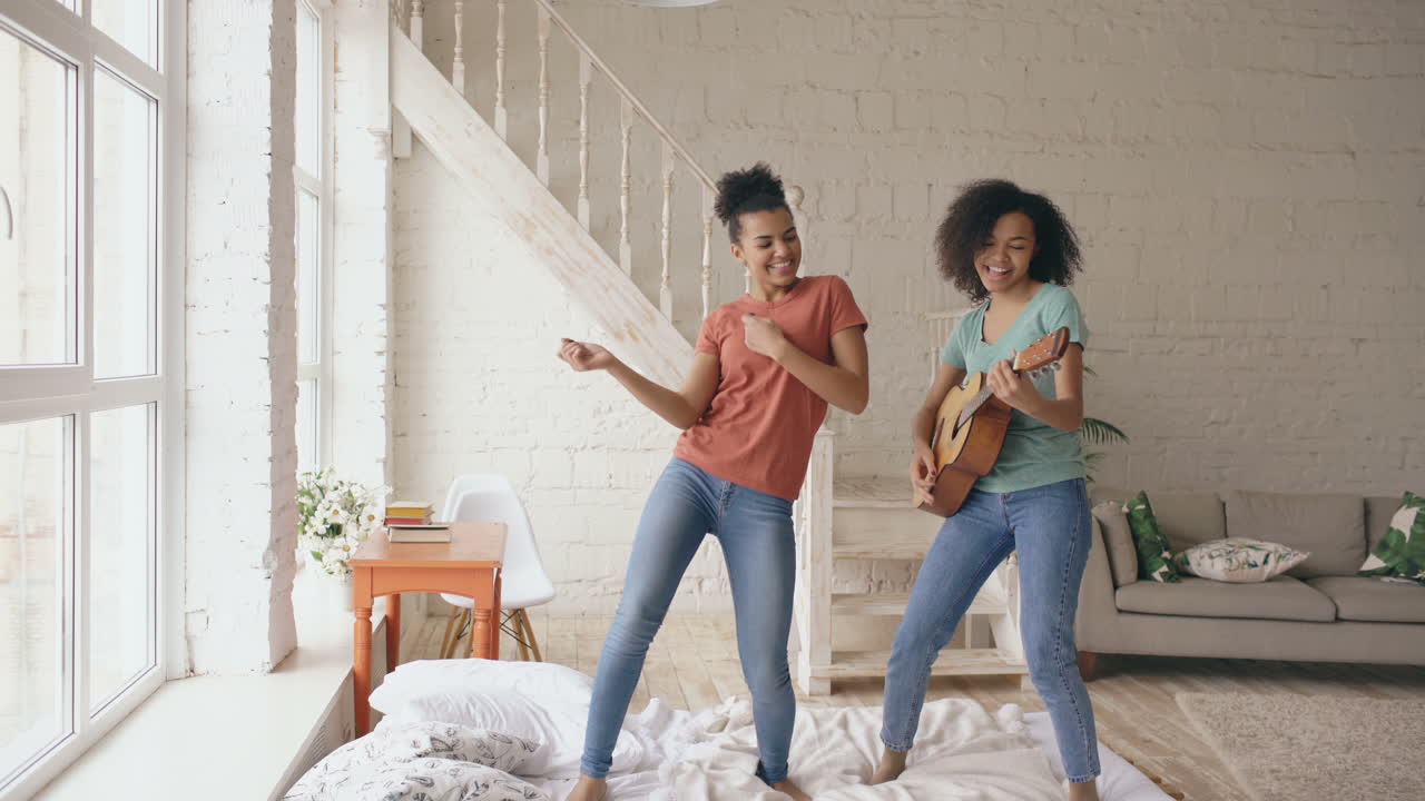 Glückliche Teenagerinnen tanzen und spielen Gitarre auf einem Bett