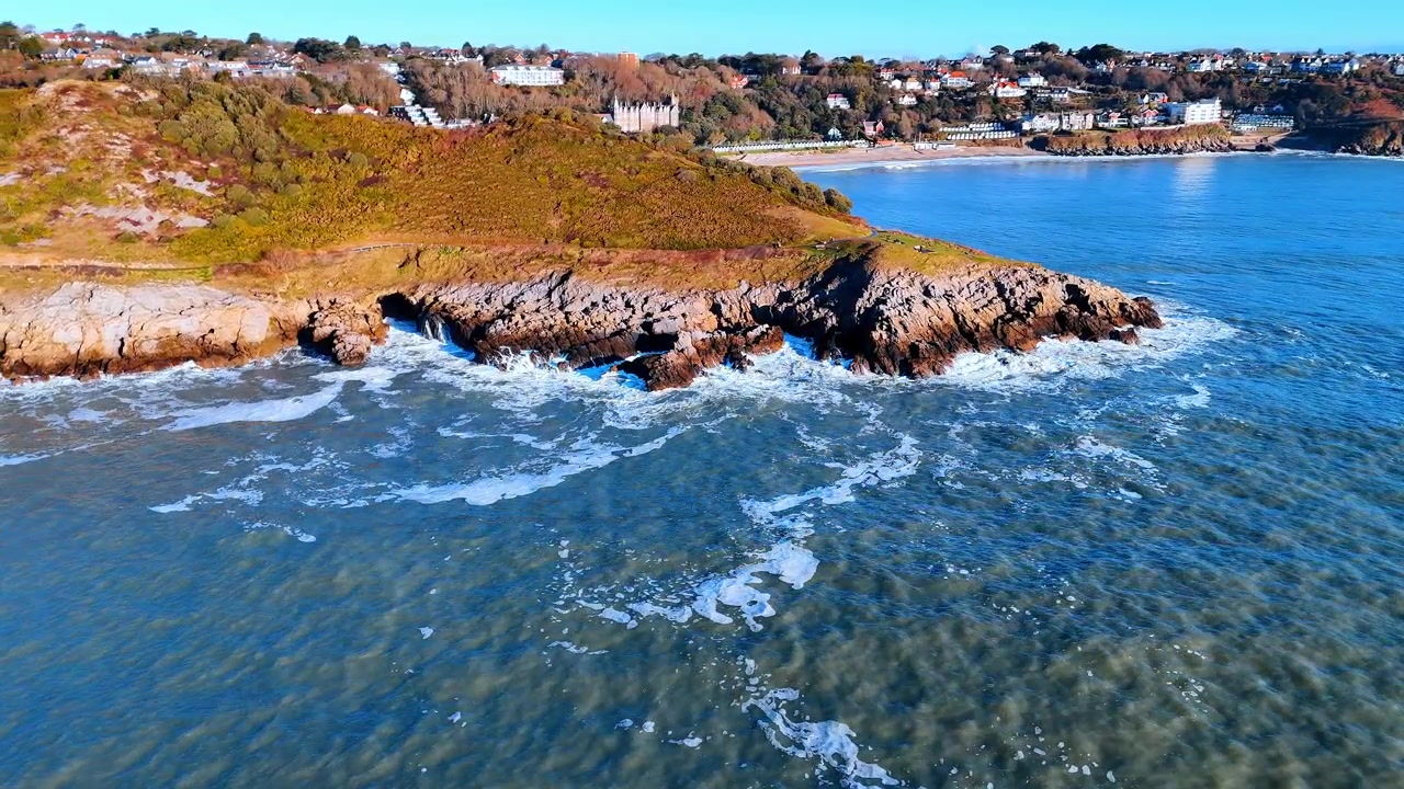 Flygpanorama över Walesiska kusten med golfbana och udde. En bred udde med en grön golfbana och rostfärgad ljunghed möter havet när kustlinjen kröker sig mot avlägsna klippor.