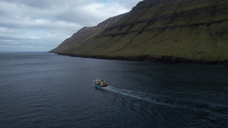 aerial view in a circle of a fishing boat that sails through a fjord in the faroe islands and where the great mountains can be seen